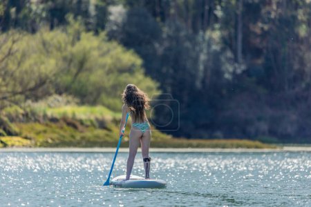 teenage girl seen from behind, standing on her paddleboard and navigating through a river surrounded by lush nature, enjoying outdoor water sports and leisure activities