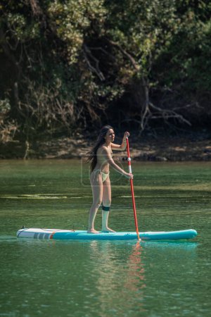 mujer latina joven vertical en bikini disfrutando de una excursión de stand up paddleboarding en el río, sumergiéndose en la naturaleza en un día de verano