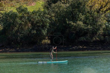 Side view of a young multicultural woman, wearing a bikini, enjoying stand up paddle on the river surrounded by nature, enjoying outdoor sports and leisure activities