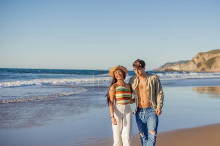 copyspace multicultural Latina couple walking along the beach shore, their faces illuminated by the warm glow of the sunset. Their smiles radiate joy and contentment as they enjoy a peaceful stroll