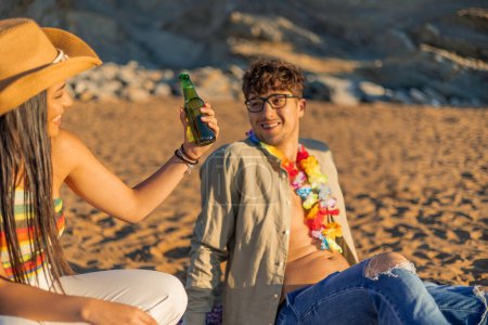 Téléchargez les photos : Une femme hispanique offre une bière à son petit ami alors qu'ils sont assis ensemble sur la plage de sable fin pendant un coucher de soleil d'été. Leur moment intime est rempli de chaleur et la beauté tranquille de la mer du soir - en image libre de droit