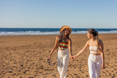 Zwei hispanische multikulturelle lesbische Mädchen gehen Hand in Hand und drücken ihre Liebe aus, während sie an einem sonnigen Sommertag am Strand spazieren und die Schönheit von LGBTQ + -Beziehungen inmitten der Küstenlandschaft präsentieren.