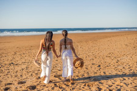 multikulturelles lesbisches Paar aus Latina, das an einem Sommertag Hand in Hand am Strand entlang läuft, mit dem Rücken zur Kamera, während sie ihre Liebe und Zuneigung durch ihre ineinander verschlungenen Finger ausdrücken
