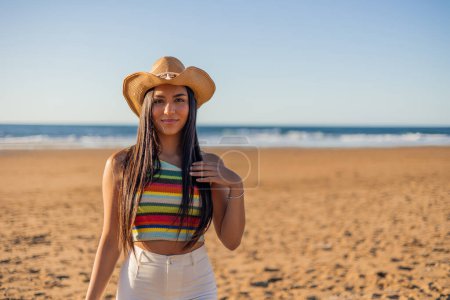multicultural Latin woman striking a pose on the beach, adorned with a stylish hat, basking in the warm glow of a sunny summer day.