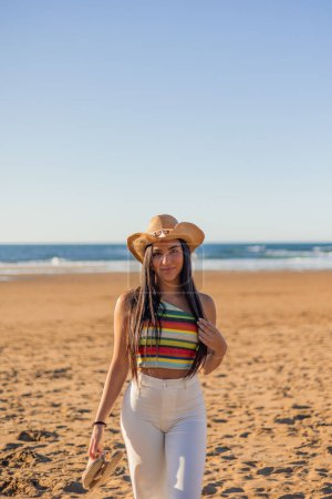 vertikale multikulturelle Schönheit als Latina-Frau posiert am sonnigen Strand, ihr stylischer Sonnenhut verleiht der lebendigen Szene einen Hauch von Eleganz.