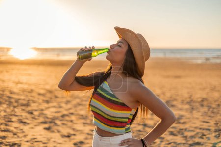 multicultural Latin girl savors a beer bottle on the beach, soaking in the serene ambiance of summer sunset. Her relaxed demeanor and the tranquil seaside setting create a perfect moment of relaxation