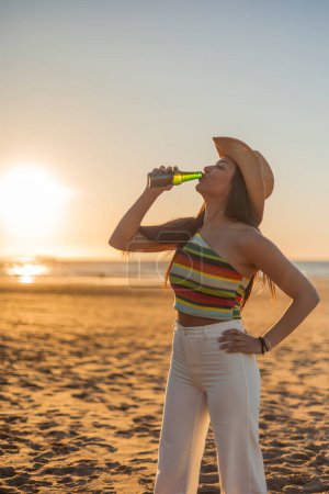 vertical carefree evening on the beach as a young Latina woman savors a refreshing beer against the backdrop of a stunning summer sunset, With the golden glow of the sun dipping below the horizon