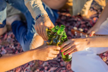 closeup shot captures the hands of three Latin friends raising beer bottles in a toast on the beach during a summer sunset. The image portrays the camaraderie and joy of friendship