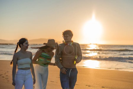 drei multikulturelle Freunde spazieren am Strand entlang und teilen Lachen und Freude, während sie die friedliche Atmosphäre des Sonnenuntergangs genießen