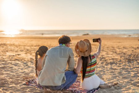 Three multicultural friends are seen from behind as they take a selfie with a cellphone on the beach, gazing at the sunset over the sea. The image captures the camaraderie and joy of friendship
