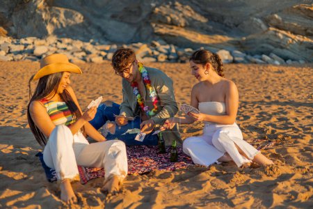 Téléchargez les photos : Trois amis hispaniques se rassemblent sur la plage, jouent aux cartes et s'amusent au coucher du soleil d'été. Leurs rires et leur plaisir remplissent l'air, créant une soirée mémorable de bord de mer - en image libre de droit