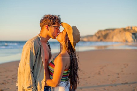 Un momento romántico y apasionado capturado mientras una pareja hispana multicultural se besa profundamente en la playa durante una hermosa puesta de sol de verano, con el mar al fondo