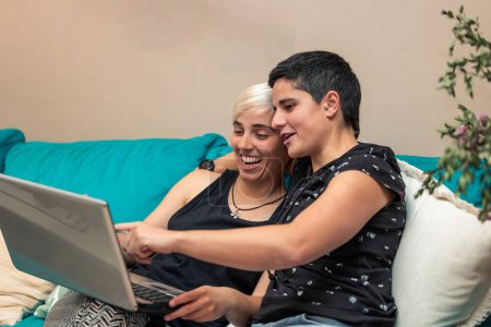 Dos mujeres, una pareja de lesbianas, sentadas juntas en el sofá, navegando tranquilamente por Internet en su portátil. Estilo de vida moderno y concepto de tecnología