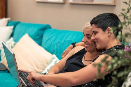 Dos mujeres, una pareja homosexual, abrazándose cariñosamente mientras se divierten con un portátil en el sofá de casa. Ocio moderno y concepto de estilo de vida LGBTQ +