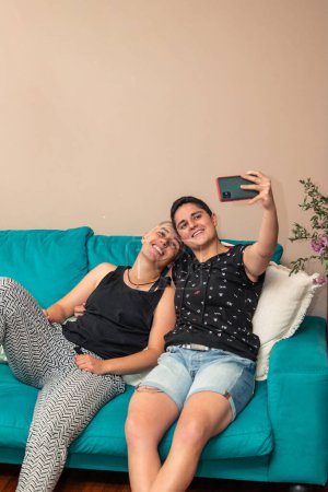 vertical Pareja de chicas homosexuales relajadas sentadas en su sofá casero tomando una selfie con teléfono celular. Estilo de vida moderno y concepto de amor