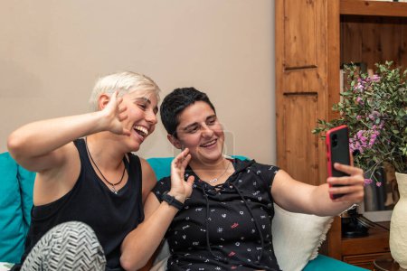 Dos mujeres, una pareja de lesbianas, felizmente hacen una videollamada con sus amigos usando un teléfono inteligente desde la comodidad de su hogar. Concepto moderno de socialización y estilo de vida LGBTQ +