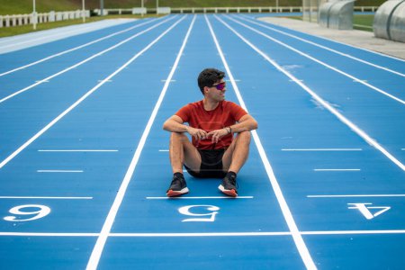 Der mit Sonnenbrille ausgestattete männliche Läufer liegt nach einem langen Rennen auf der Ziellinie einer blauen Leichtathletikbahn am Boden. fängt den Moment der Entspannung und Erleichterung ein, wenn sich der Sportler erholt