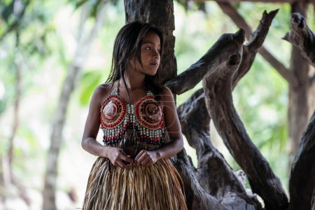 Das junge indigene Mädchen der Wayuri in traditioneller Kleidung blickt nach rechts, während es sich im ecuadorianischen Amazonas-Dschungel an einen Baum lehnt. fängt ihre Verbindung mit der natürlichen Umwelt ein