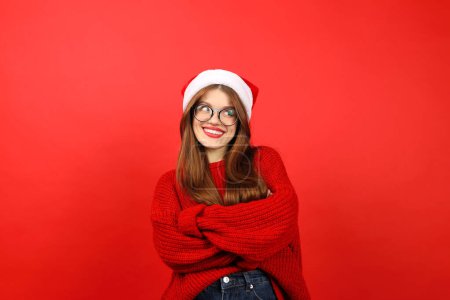 Téléchargez les photos : Doutant femme gaie dans le chapeau de Père Noël sur fond rouge, pensant confusément aux cadeaux de Noël. - en image libre de droit
