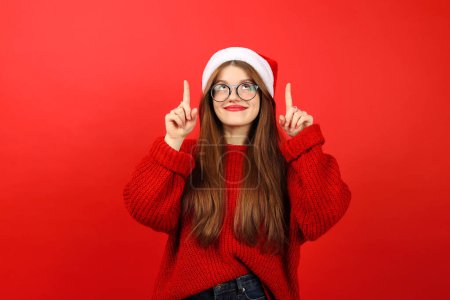 Foto de Chica alegre en un sombrero de santa y un suéter festivo apunta hacia arriba con las manos sobre un fondo rojo. Lugar de publicidad o venta. - Imagen libre de derechos