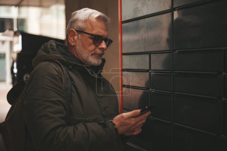 Photo for Senior man picking up a package from a post machine late at night after work. Express delivery of goods from online stores. - Royalty Free Image
