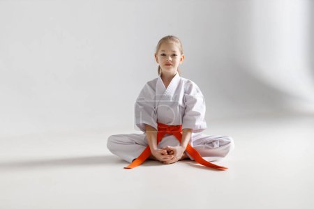 Foto de A little girl in a white kimono sits on a light background, karate. - Imagen libre de derechos