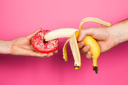 Téléchargez les photos : Erotic banana and donut in the hands as a symbol of the penis and vagina isolated on a pink background. The relationship between a man and a woman, sex and passion. - en image libre de droit