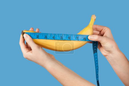 Téléchargez les photos : Measuring the size of a banana as a symbol of the male penis isolated on blue background. Big dick length. Strong erection and impotence problem. - en image libre de droit