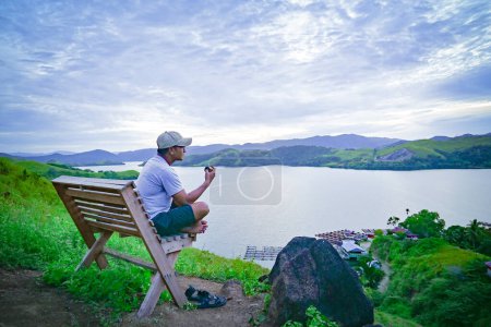 Téléchargez les photos : Un homme est assis sur un banc profitant du magnifique paysage naturel sur les rives du lac Sentani, en Papouasie depuis le sommet de la montagne - en image libre de droit