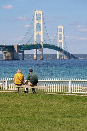 Photo for Mackinaw City, MI - September 15 2023: A couple sitting on a park bench with the Mackinaw Bridge in the distance - Royalty Free Image