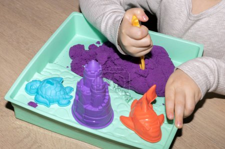 A child is using their fingers to create art with violet and magenta kinetic sand on a table. The leisure activity involves sharing gestures and thumb movements