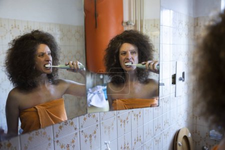 Téléchargez les photos : Mid Adult cheveux bouclés femme brossant ses dents avec une brosse électrique en face du miroir de salle de bain domestique - en image libre de droit