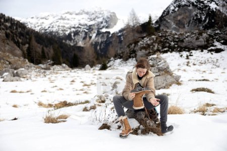 Foto de Mujer adulta quita una pequeña piedra de un zapato de senderismo en una caminata de montaña de invierno - Imagen libre de derechos