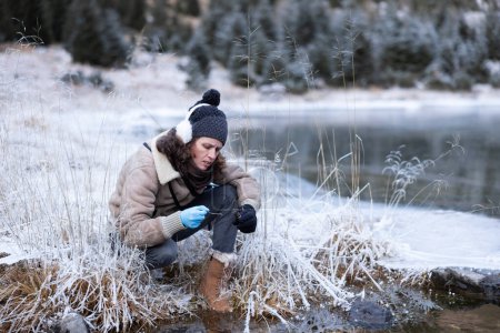 Foto de Investigadora del medio ambiente examinando la presencia de algas en cuclillas cerca de un lago congelado - Imagen libre de derechos