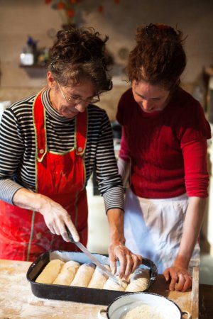 Photo for Expert Senior Woman Baker Teaching a Younger Adult Woman the Art of Hand Crafting Bread Dough - Here Giving the Final Design Touch - Royalty Free Image