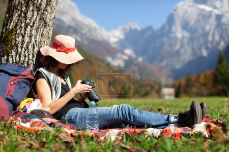 Téléchargez les photos : Femme randonneur photographe touristique examinant ses photos sur l'appareil photo tout en se reposant sous un arbre sur une belle campagne Montagnes Emplacement - en image libre de droit
