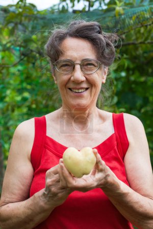 Photo for Senior Woman Smiling in Camera with in Hands Holding a Potato with heart Shape - Royalty Free Image