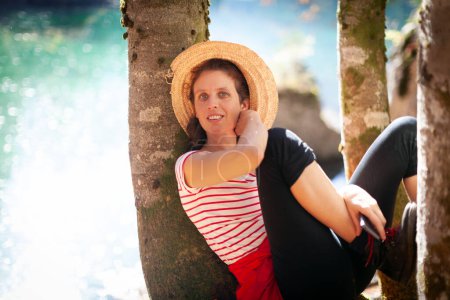 Mujer adulta relajada posando mientras está sentada en un árbol sobre el lago de montaña en la luz reflejada del rayo de sol