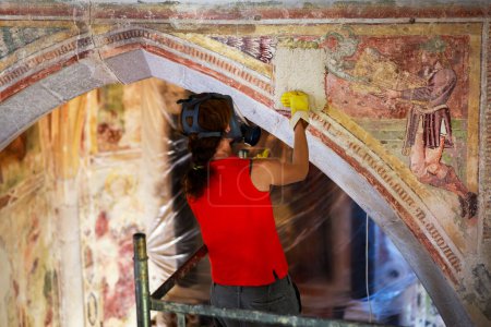 Téléchargez les photos : Processus de restauration d'applications chimiques spéciales sur un mur de voûte d'église Fresque antique par un restaurateur féminin professionnel portant un masque à gaz protecteur pendant le travail - en image libre de droit