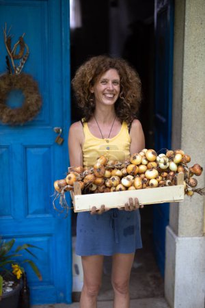 Téléchargez les photos : Joyeux caucasien souriant cheveux bouclés femme adulte agriculteur montrant ses oignons produits maison dans une caisse en bois tricoté en tresses vintage prêt à être vendu - en image libre de droit