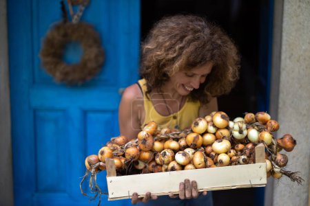 Téléchargez les photos : Femme souriante regardant vers ses oignons maison dans la caisse qu'elle tient en face de sa ferme biologique dans la campagne - en image libre de droit
