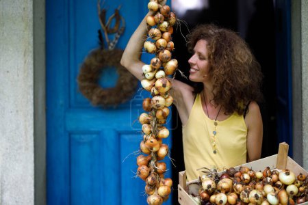 Téléchargez les photos : Jeune agricultrice adulte sereine fière de ses oignons biologiques faits maison tricotés dans une tresse traditionnelle d'oignons à l'ancienne devant sa ferme de campagne - en image libre de droit