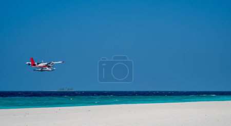 Seaplane is flying low above sea level by the sandy beach. Blue sky.