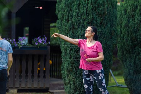 Téléchargez les photos : Une femme âgée jouant aux fléchettes dans le jardin. Elle est en plein air un jour d'été - en image libre de droit