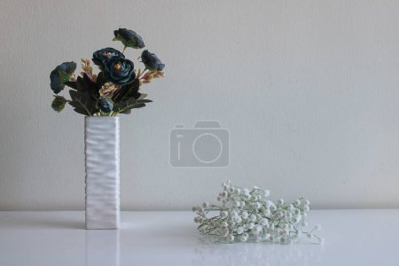 Photo for A bouquet of artificial blue flowers stands in a relief vase. Small white flowers lie nearby. Neutral background and surface - Royalty Free Image