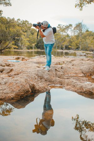 Foto de Fotgrafo de pie, tomo una foto, con su reflejo en pequeo lago y arboles de fondo - Imagen libre de derechos
