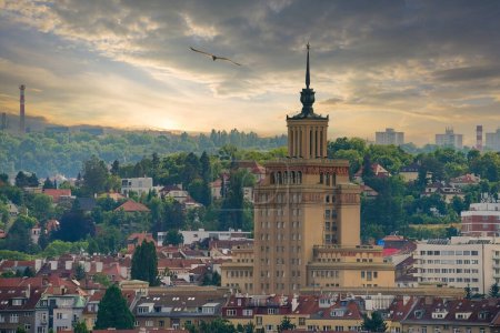 Une vue panoramique de Prague avec un bâtiment historique avec une tour et une flèche, entouré de maisons recouvertes de verdure et sur un terrain en pente, sous un ciel dramatique avec une lueur chaude.