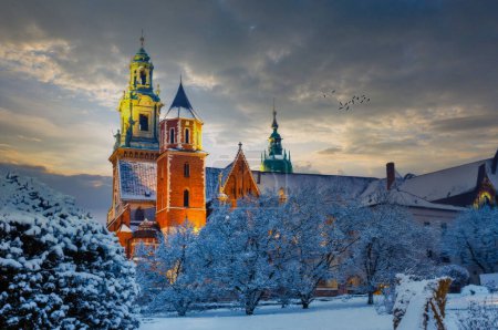 A serene winter scene in Krakow, Poland, showcasing a historic building with a greenroofed tower and gold cupola, amidst snowcovered trees at sunrise or sunset.