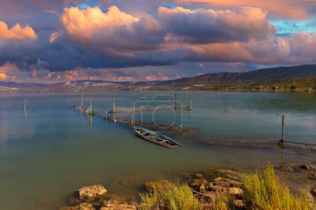 Lever ou coucher de soleil doré se reflète sur un lac serein avec un bateau submergé, entouré de collines et de nuages pelucheux, créant une scène paisible mais mélancolique.