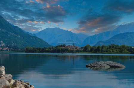 Serene lakeside dawn or dusk scene with a calm, reflective surface, a rocky outcrop, a quaint village with a church, and majestic mountains under a pastel sky, likely in Europe.